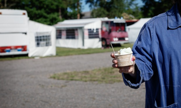 En hand som håller i glass i förgrunden, campingbilar i bakgrunden. Foto. 