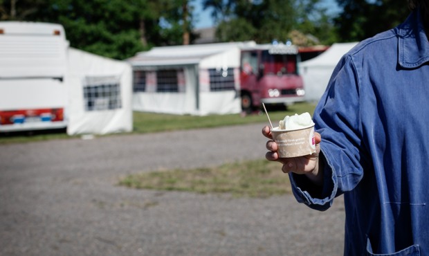 En hand som håller i glass i förgrunden, campingbilar i bakgrunden. Foto.