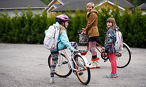 Foto på vuxen och två barn med cyklar på en gata i ett villaområde.