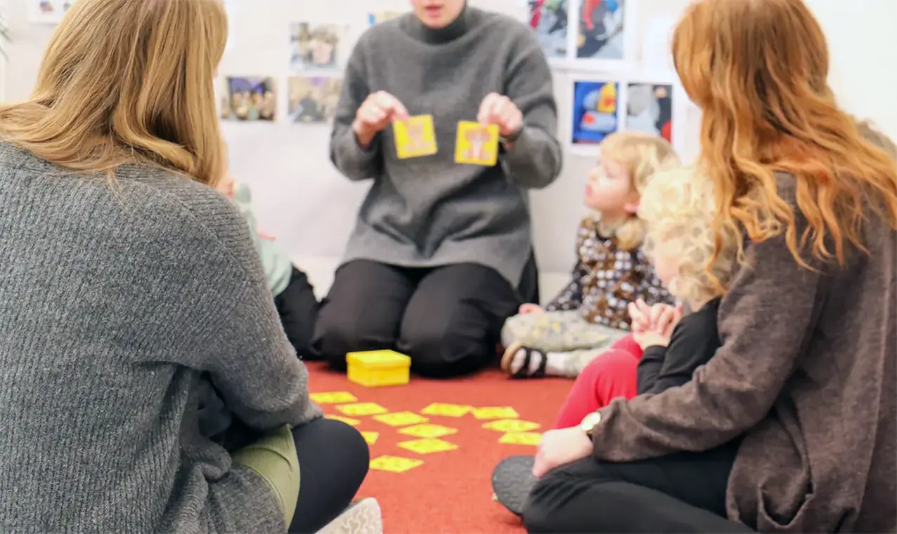 Tre vuxna och några barn sitter på en matta på golvet i förskolemiljö. En av de vuxna håller upp gula kort med bilder på. 